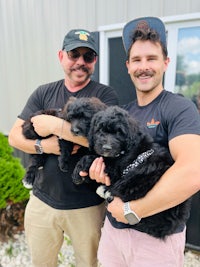 two men holding black puppies in front of a building